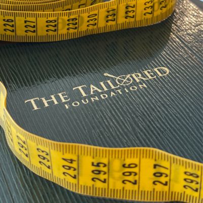 Yellow tailor's tape measure on dark suiting cloth embossed with The Tailored Foundation logo