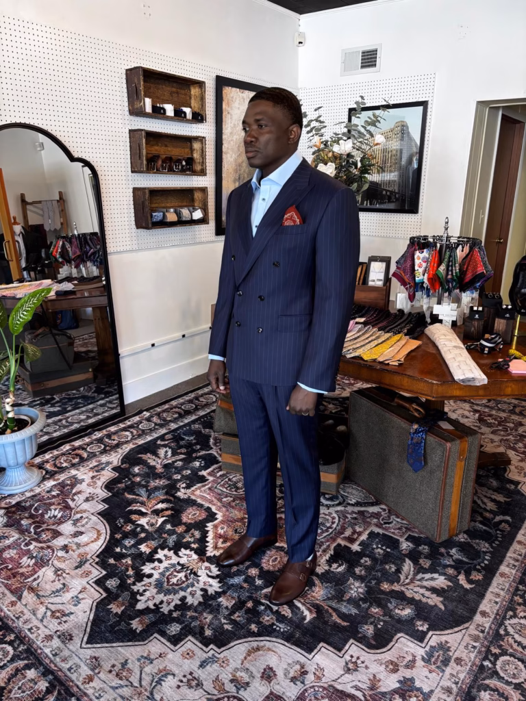 WhatsApp Image 2026-05-22 at 03.53.37 (1) Man wearing a navy pinstripe bespoke suit inside a tailoring studio