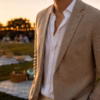Close-up of a man wearing a tan sport coat over an open-collar white shirt at an outdoor golden-hour picnic with blurred string lights and blankets in the background.