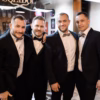 Front view of groom and his friends wearing in black festive outfits embracing each other while posing and looking at camera in barber shop in wed day