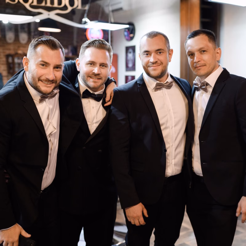 Front view of groom and his friends wearing in black festive outfits embracing each other while posing and looking at camera in barber shop in wed day